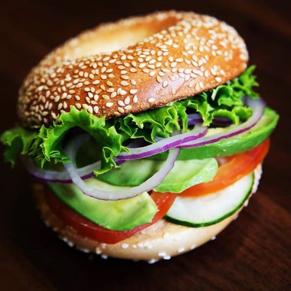 A sesame bagel sandwich with lettuce, red onion, avocado, tomato, and cucumber on a dark surface.