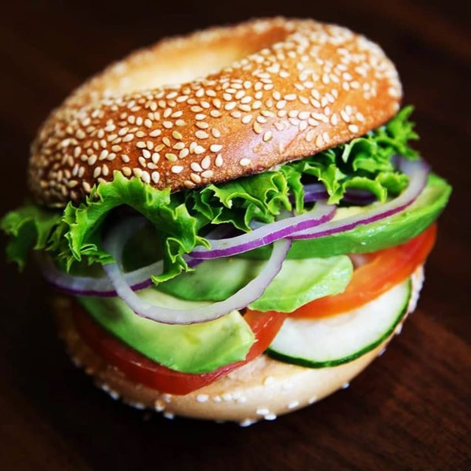 A sesame bagel sandwich with lettuce, red onion, avocado, tomato, and cucumber on a dark surface.